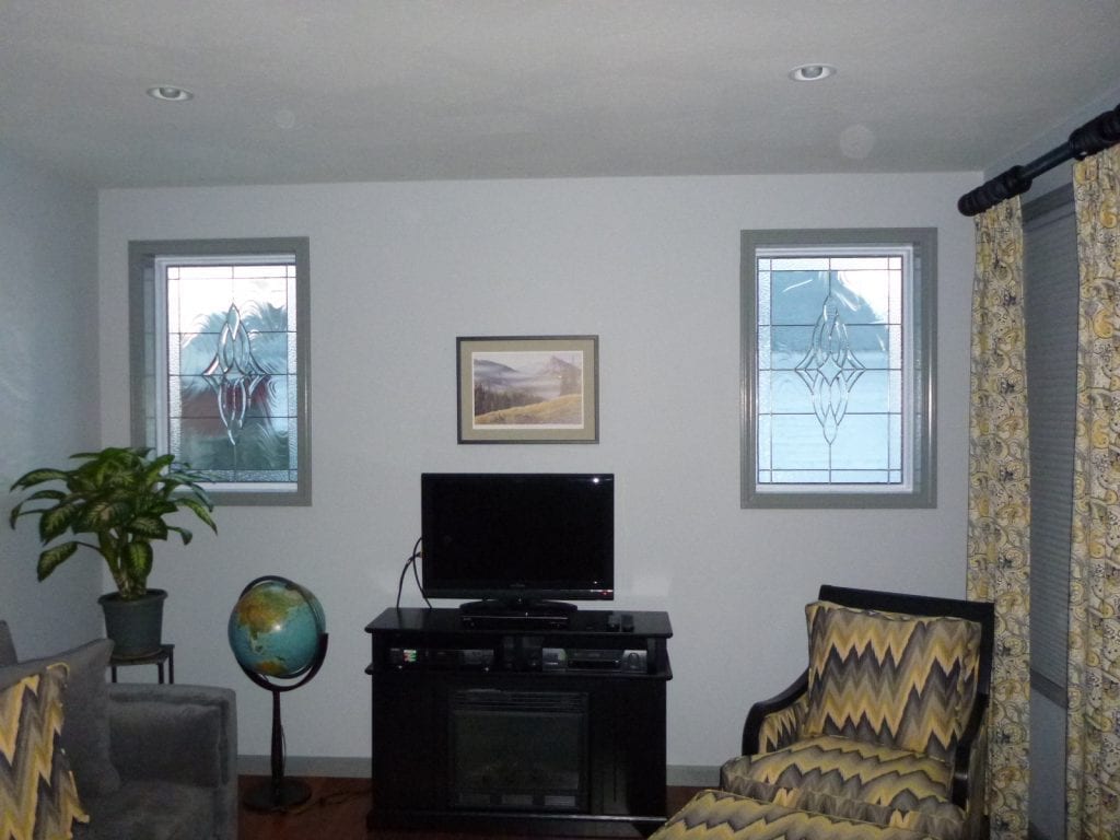 Two rectangular leaded stained glass panels installed in side-by-side windows within a living room. Each panel features a unique, symmetrical abstract design created with clear, textured, and beveled glass pieces. The designs include curved lines and decorative motifs, providing privacy while allowing light through.