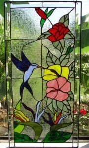 A vibrant rectangular stained glass panel featuring a blue hummingbird hovering near yellow and pink hibiscus flowers, with red roses and orange bird of paradise blooms, all set against a clear textured background.