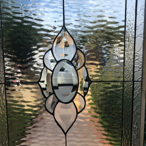 A rectangular leaded stained glass panel with a central geometric medallion formed from various clear beveled glass pieces. The surrounding background glass is a rippled clear water glass texture, which distorts the view of trees and sky outside. The far right edge of the panel features a more pronounced textured clear glass, possibly glue chip or frosted, adding to the privacy.