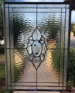 A rectangular clear leaded stained glass panel with a central abstract floral or petal-like design made of clear beveled glass. The surrounding glass features distinct sections of clear ripple (water) and glue chip textures, all encased in a metal frame.