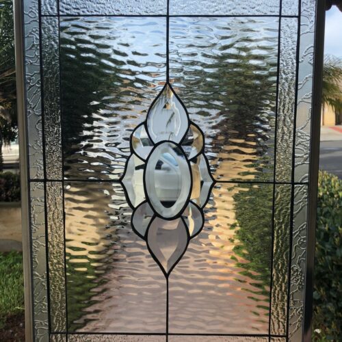 A rectangular clear leaded stained glass panel with a central abstract floral or petal-like design made of clear beveled glass. The surrounding glass features distinct sections of clear ripple (water) and glue chip textures, all encased in a metal frame.