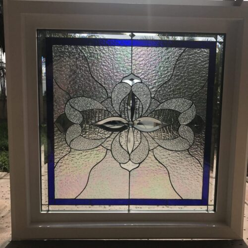 A square stained glass panel encased in a robust white frame, featuring a central abstract floral or butterfly-like design. The leaded glass pattern uses clear, highly textured, and iridescent glass with sparkling bevels, all framed by a distinct deep blue glass border.