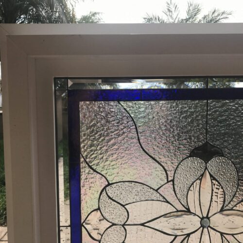 Rectangular leaded stained glass panel with a central clear textured floral design, surrounded by various clear textured glass, and accented with a deep blue border on the left and top. The panel is set in a white frame, overlooking a green yard and palm trees.