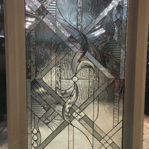 A rectangular clear stained glass panel, showcasing an intricate abstract and geometric design composed of various clear textured, beveled, and heavily textured glass sections. The central motif features elegant swirling clear glass, surrounded by a dynamic mosaic of horizontal, vertical, and diagonal patterns. Many sections include sparkling, faceted glass elements that brilliantly refract light. The panel is set in a light-colored frame, with natural light from outdoors illuminating the textures and creating bright reflections.
