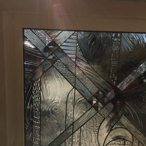 A rectangular clear stained glass panel installed in a light-colored frame, featuring an intricate abstract geometric design composed of diverse textured glass, beveled accents, and lead lines. Sunlight shining through creates prism effects and highlights the various clear glass patterns, including wavy, striped, pebbled, and diamond-shaped bevels, with faint palm fronds and sky visible outside.