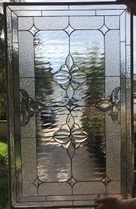 A rectangular clear stained glass panel with a symmetrical design. It features a central vertical arrangement of two large, intricate beveled glass floral medallions. Two smaller, partial floral motifs extend horizontally from the sides. The main background sections are clear waterglass, framed by textured clear hammered glass borders and thin clear beveled pieces. All glass pieces are joined by dark lead came.