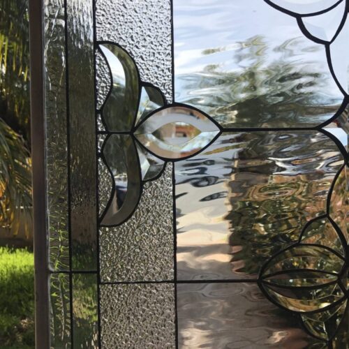 Close-up of a clear leaded stained glass panel featuring a central floral or leaf-like design made with clear beveled glass, flanked by vertical sections of clear textured glass, including ripple and glue chip patterns. The outdoor view through the glass shows blurred green foliage and sunlight.