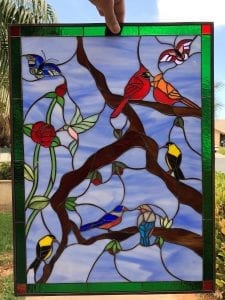 A rectangular stained glass panel, crafted with copper foil, showcasing a vibrant nature scene. Multiple colorful songbirds, including red cardinals, yellow orioles, and bluebirds, perch on brown tree branches alongside red roses, a hummingbird, and two butterflies, all set against a light blue sky. The panel is bordered with green and red accent glass.