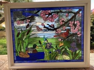 A rectangular stained glass panel depicting a vibrant spring nature scene. A blue bird and a red cardinal perch on branches adorned with pink cherry blossoms. In the foreground, two ducks swim in a blue pond, surrounded by green cattails and purple irises. A hummingbird hovers over the flowers, and a butterfly is visible. The detailed panel is set within a clean white frame, showcasing varied glass textures and lead lines.