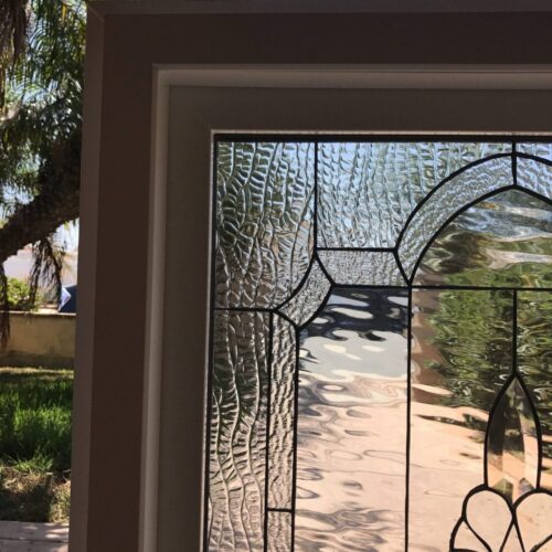 A detailed view of a clear stained glass sidelight panel, featuring a mix of textured and beveled glass pieces creating an elegant geometric design with a subtle floral accent at the bottom right. The panel is set within a light-colored frame, with blurred palm trees and a sunny sky visible outside, reflecting off the glass.