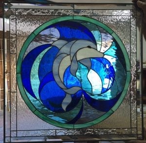 A square leaded stained glass panel featuring a central circular design. A graceful gray and white dolphin is depicted swimming amidst swirling waves of deep blue, light blue, and aqua stained glass. The outer square border consists of clear, highly textured 'water glass' that resembles ripples.