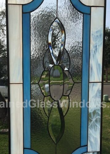 A tall rectangular leaded stained glass panel. The central design features clear, textured glass, with a vertical column of clear beveled glass elements resembling three stacked abstract flowers or elongated petals. This centerpiece is framed by a rich turquoise blue glass border with rounded corners at the top and bottom, which is then surrounded by an outer border of streaky white opalescent glass. The lead lines are dark, and the panel is viewed outdoors against a natural background.