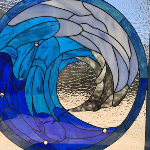 A round stained glass panel depicting a stylized ocean wave in various shades of deep blue, sky blue, and white, with clear textured glass and three small clear jewels, set against a background of clear textured glass.
