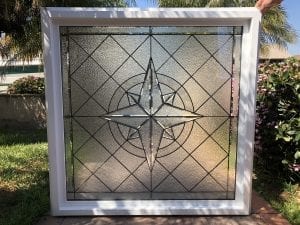 A square insulated window with a white vinyl frame, featuring a leaded stained glass panel. The design showcases a prominent central compass rose or starburst made from clear beveled glass, surrounded by a background of clear textured glass with a geometric grid and concentric circles, against an outdoor setting.