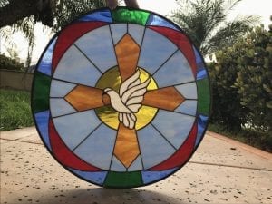 A circular leaded stained glass panel featuring a white dove with outstretched wings at its center, perched on an amber-colored cross. The cross is set against a golden yellow halo, surrounded by light blue radiating glass sections, and finished with an outer border of red, blue, and green segmented glass.