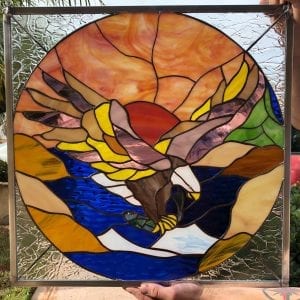 A square, leaded stained glass panel featuring a circular central design. The design depicts a majestic brown, white, and yellow bald eagle with outstretched wings, soaring downwards towards dark blue water. The eagle clutches a silver-blue fish in its talons. Behind the eagle, a vibrant red, orange, and yellow sunrise or sunset glows over green and brown mountains. The outer square areas of the panel, surrounding the circular scene, use clear, textured stained glass.