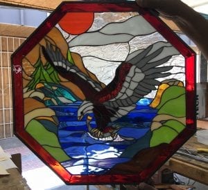 An octagonal stained glass panel depicting a powerful bald eagle with white head and tail, dark brown body, and grey wings, actively catching a silver fish from a vibrant blue body of water. The scene features green and brown mountains, an orange sun, and white clouds, all framed by a deep red border.