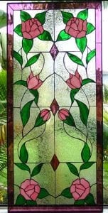 A rectangular stained glass window panel featuring a symmetrical floral design. The panel has a dark purple border. Inside, two pink roses bloom at the top corners and two at the bottom corners. In the middle section, two pink rose buds appear on each side, with diamond-shaped accents in the center. Vibrant green leaves and flowing black lead lines connect the floral elements against a textured clear glass background.