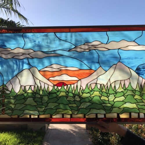 Rectangular leaded stained glass panel featuring a scenic mountain landscape at sunset, with a vibrant orange and red sun partially obscured by white peaks, lush green pine trees in the foreground, and a blue sky with soft white clouds.