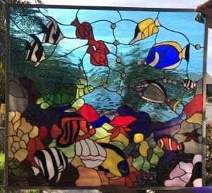 A rectangular leaded stained glass panel depicting a lively underwater coral reef scene. It features a variety of colorful tropical fish including black and white striped angelfish, a bright blue surgeonfish, a clownfish, and others in red, yellow, and orange. A green sea turtle swims among intricately detailed corals and anemones in shades of red, orange, pink, and brown. The upper portion shows rippling blue water, conveying depth and movement.