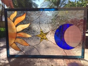 A rectangular stained glass panel depicts a celestial scene: a radiant orange and yellow sun on the left, a vibrant blue crescent moon paired with a clear reeded glass circle on the right, and a golden five-pointed star in the middle. The background is clear, highly textured crackle glass, all encased in a dark metal frame.