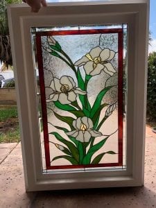 A rectangular stained glass window panel depicting white iris flowers with green leaves and gold accents, framed by a red border and clear textured glass, set within a white insulated window frame.