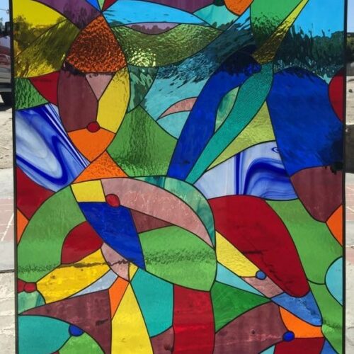 A vibrant rectangular stained glass panel featuring a complex, abstract design composed of numerous irregular, interconnected shapes. The piece is rich with diverse colors including bright blues, greens, reds, yellows, oranges, and purples. Many glass segments display varied textures like streaky, rippled, and clear smooth patterns, all framed by a slim black border. It is held up against a bright outdoor sky, enhancing its luminosity.