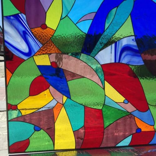 A rectangular stained glass panel featuring a vibrant, abstract geometric design. The pattern is composed of numerous leaded glass segments in a rich array of colors including deep reds, blues, greens, yellows, purples, oranges, and turquoise. Various glass textures are visible, some smooth, some rippled or mottled, creating a dynamic play of light.