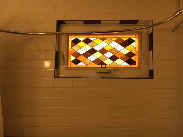 A rectangular stained glass window, set in a white vinyl frame, features a diamond or harlequin pattern using amber, yellow, brown, and white translucent glass panes. The window is installed in a cream subway-tiled bathroom wall, above a shower area, with a silver curved shower rod visible. A black and white checkered border surrounds the window opening.