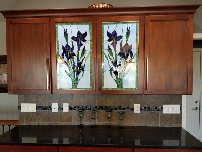 A pair of custom stained glass panels featuring vibrant purple and yellow iris flowers with green foliage, integrated into light brown wooden kitchen cabinet doors, positioned above a dark granite countertop and a tiled kitchen backsplash with mosaic accents.