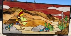 A colorful rectangular stained glass panel depicting a vibrant Southwest desert landscape. A stylized Kokopelli figure in light green plays a flute in the foreground, accompanied by a blue and green striped lizard, two small blue and red birds, and desert plants like cacti and a white yucca bloom. The background shows rolling hills in shades of red, brown, and tan.