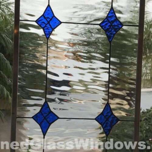 A rectangular leaded stained glass panel with a dark metal frame, featuring a clear, heavily rippled glass background. Eight vibrant cobalt blue diamond-shaped glass pieces are arranged symmetrically in two vertical columns of four, connected by dark lead lines. The panel is held outdoors against a blurred background of green foliage and a light sky.