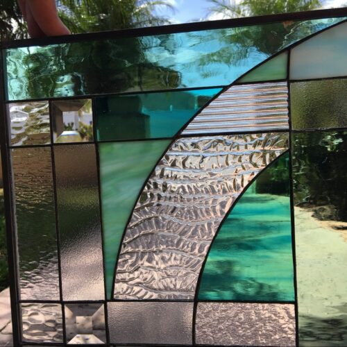A rectangular stained glass panel featuring an abstract geometric design with shades of green and teal art glass, and various clear textured glass sections. A prominent S-shaped curve of clear, heavily textured ripple glass sweeps through the center, bordered by smooth and streaky colored glass pieces, and includes two clear square bevels in the bottom left, all enclosed in a dark frame.