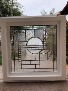 A rectangular, white-framed leaded stained glass panel showcasing an intricate abstract geometric design. The panel is crafted from various clear textured glass types, including fluted, hammered, and seedy, with a prominent central circular motif.