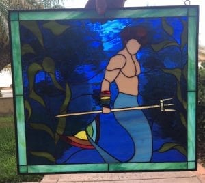 A rectangular stained glass panel featuring a muscular merman with brown hair and a light blue tail, holding a golden trident. The background is a vibrant, textured dark blue representing water, adorned with flowing green seaweed. The panel is framed by a lighter green border and has black lead lines.