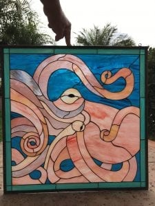 A rectangular stained glass art panel featuring a large, stylized octopus in various shades of pink, peach, and cream with dynamic, swirling tentacles. The octopus is set against a vibrant, textured blue glass background representing water. The panel has a slim teal border and a dark metal frame with a visible hanging loop.