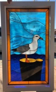 A rectangular stained glass window panel in a white frame, featuring a white and gray seagull with an orange beak and feet, standing on two dark pilings with yellow tops. The background depicts wavy ocean water and sky, crafted from various shades of textured blue art glass. An inner border of amber-orange glass frames the central scene.