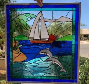 A vibrant rectangular leaded stained glass panel depicting a sailboat with white sails on deep blue water, with two gray dolphins leaping from teal waves. An orange sun sets behind the boat, casting rays, with green coastal landscapes on either side. The panel is bordered with broad royal blue and narrower teal stained glass.