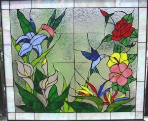 A rectangular stained glass panel depicting a vibrant tropical garden scene. Two hummingbirds, one pink and one blue, are shown feeding from an array of colorful flowers. The arrangement includes blue lilies, white calla lilies, red roses, yellow and pink hibiscus, and orange and blue bird of paradise flowers, all set against a clear textured background with a subtle clear opalescent border.