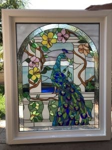 A rectangular stained glass window panel in a white frame, featuring an arched design. The central image is a vibrant peacock with shimmering blue, green, and purple feathers, perched on a white balustrade. The peacock is surrounded by a lush garden scene with green vines, brown branches, and colorful pink and yellow flowers. The background includes light blue and white glass, with some clear textured glass sections.