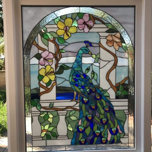 A rectangular stained glass window panel in a white frame, featuring an arched design. The central image is a vibrant peacock with shimmering blue, green, and purple feathers, perched on a white balustrade. The peacock is surrounded by a lush garden scene with green vines, brown branches, and colorful pink and yellow flowers. The background includes light blue and white glass, with some clear textured glass sections.