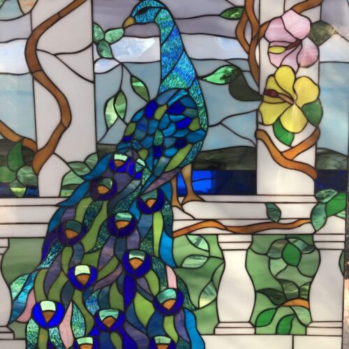 A vibrant rectangular stained glass panel featuring a majestic peacock with iridescent blue and green plumage, perched on a white architectural balustrade. Lush green foliage and colorful pink and yellow flowers on winding brown vines frame the scene, set against a background of soft blue sky and water.
