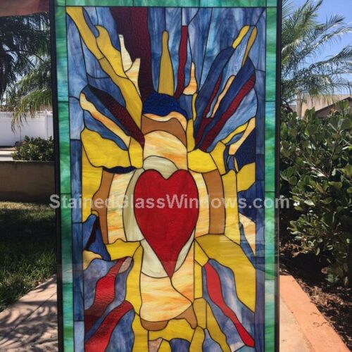 A rectangular stained glass panel featuring a prominent red heart at its center, surrounded by an abstract composition of radiating yellow, orange, blue, and deep red glass, all contained within a dark leaded frame.