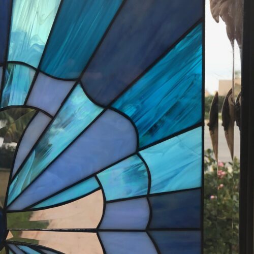 A close-up of a vibrant stained glass window panel featuring a radial, fan-like geometric design. The panel showcases diverse shades of blue and teal art glass, including streaky, opalescent, and clear beveled textures, outlined by dark lead lines.