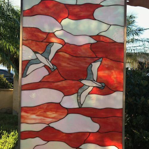 A rectangular stained glass panel depicting two seagulls flying across a vibrant sunset or sunrise. The sky and water are composed of swirling red and orange streaky glass, with white and iridescent glass forming soft clouds. Two stylized seagulls with white bodies and light grey-blue wings are in flight. A dark red circular sun or its reflection is at the bottom, all framed in metal.