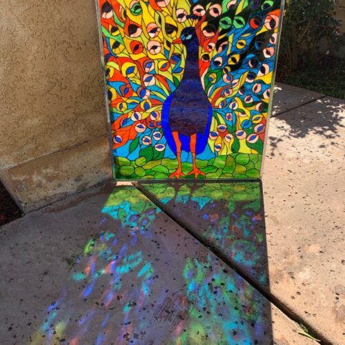 Vibrant stained glass panel featuring a majestic peacock with its fanned tail in shades of blue, yellow, green, orange, and red, casting colorful light reflections onto the ground.
