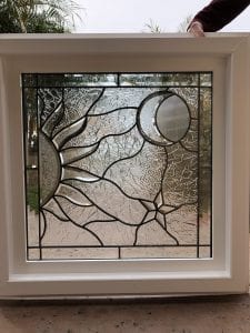 A rectangular leaded stained glass panel featuring a celestial design with a sun, a crescent moon with reeded glass, and a star. The panel uses clear, textured, and beveled glass set within a white frame.