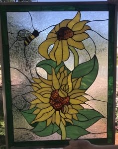 A rectangular leaded stained glass panel depicting two vibrant yellow sunflowers with brown centers and green leaves. A black and yellow bee flies towards the top sunflower. The background is made of clear, highly textured glass, and the panel is framed by a dark green glass border.