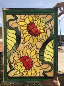 A rectangular stained glass panel showcasing two large, vibrant sunflowers. The sunflowers feature golden yellow petals and textured red-orange centers. Dark green leaves and winding stems intertwine around the flowers, set against a background of varied light cream and pale yellow glass. The entire panel is framed by a solid dark green stained glass border and a metallic outer frame, held up against a bright blue sky.