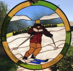 A circular stained glass suncatcher featuring a skier in a vibrant red jacket and brown pants, gliding down a white snowy mountain slope on blue skis with poles. The background includes blue sky, textured white mountains, and green foothills with dark trees, all encircled by a segmented amber and green border.